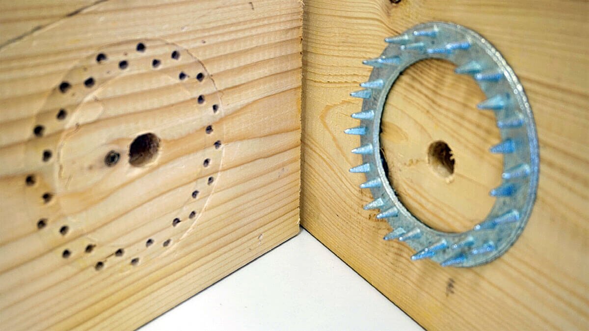 Zdjęcie przedstawia dwa drewniane elementy znajdujące się prostopadle do siebie i tworzące kąt prosty. Na elemencie po prawej stronie znajduje sie pierścień Geka, czyli stalowy okrąg wykończony ostrymi kolcami, w środku okręgu, w drewnianym elemencie znajduje się otwór. Na drewnianym elemencie po lewej stronie widoczne są niewielkie otwory tworzące okrąg, w środku którego znajduje się większy otwór.