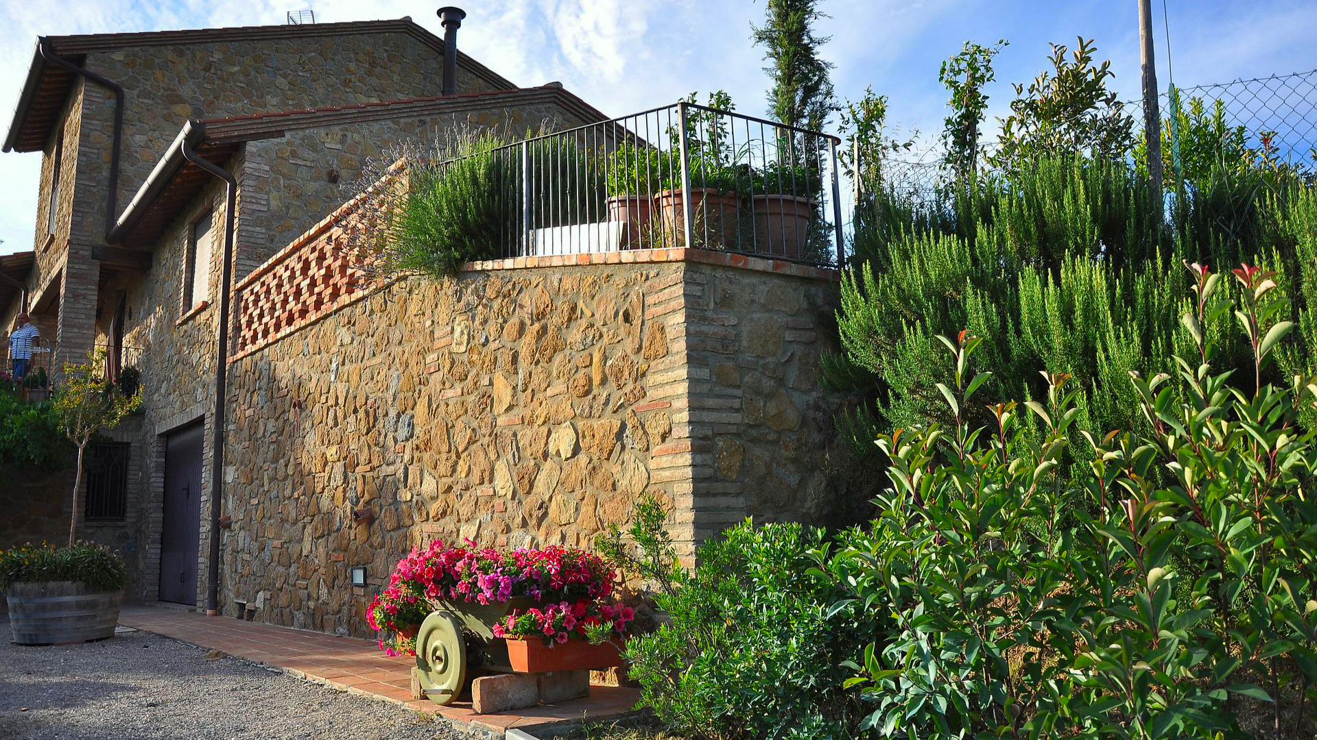 Zdjęcie przedstawia piętrowy dom zbudowany z kamienia. Przed domem widoczne są krzewy i kwiaty.