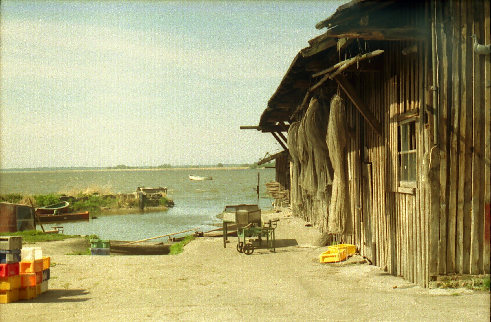 Kliknij, aby powiększyć. Ilustracja przedstawia budynek stojący przy brzegu. Znajduje się po prawej stronie. Jest zbudowany z drewna. Przy ścianie zawieszone są sieci rybackie. Przy budynku stoją skrzynie. Na jeziorze znajdują się łódki - dwie po lewej stronie w zatoczce przy brzegu, jedna na środku, w oddali, kilka‑kilkanaście metrów od brzegu.