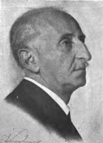 Czarno‑biała fotografia przedstawia popiersie mężczyzny w średnim wieku, ujęte w półprofilu. Ma rzadkie, siwe włosy zaczesane do tyłu z widocznymi zakolami, duży i wydatny nos, pod nim niewielki wąsik oraz małe oczy. Jest ubrany w garnitur i białą koszulę.