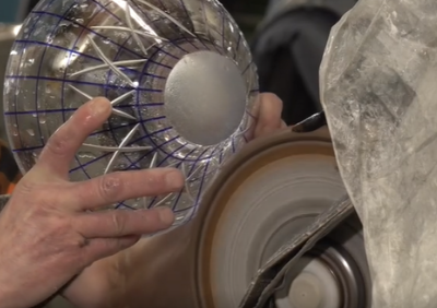 Ilustracja przedstawia szklaną miskę z narysowanym na niej wzorem. Mężczyzna trzyma ją w dłoniach nad paskiem szlifierskim. Pasek zamontowany jest na obracającym się szybko walcu. 