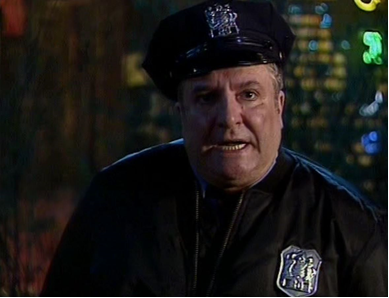 Zdjęcie przedstawia popiersie mężczyzny w stroju amerykańskiego policjanta. Mężczyzna jest tęgi, ma pełną twarz. Ma częściowo otwarte usta. Na głowie ma czapkę policyjną, ubrany jest w mundur. Na jego piersi znajduje się metalowa odznaka.