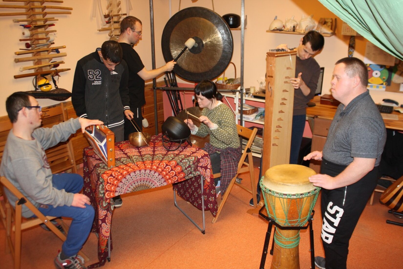 Fotografia ukazuje sześć osób w pracowni muzycznej, w której widać różne instrumenty. Cztery osoby stoją, dotykając instrumentów – są to mężczyźni. Jeden z nich jest krępej budowy, ma krótką szyję oraz ciemne włosy. Pozostali trzej są szczupli i także mają ciemne włosy. Dwie siedzące postacie to mężczyzna w niebieskich dresowych spodniach oraz szarej bluzie oraz kobieta ze spiętymi, czarnymi włosami i jasną skórą. W pomieszczeniu widoczny jest stół, przy którego końcach siedzą. Jest on zasnuty kolorowym obrusem, umieszczono na nim instrumenty. Na ścianach są półki lub uchwyty trzymające przedmioty przypominające flety, a pod nimi – meble. Podłoga jest pomarańczowa. 