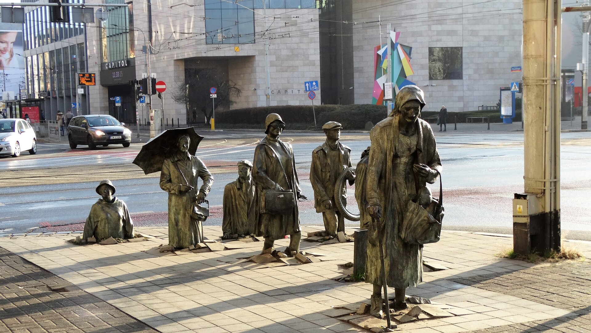 Zdjęcie przedstawia pomnik autorstwa Jerzego Kaliny, pod tytułem „Pomnik anonimowego przechodnia”. Dzieło przedstawia ludzi, postacie z brązu naturalnej wielkości. Na fotografii ukazanych jest siedem postaci. Na pierwszym planie można wyróżnić starszą kobietę z laską oraz zakupami. Tuż za nią znajduje się fragment kolejnej postaci. Na drugim planie, za kobietą na środku znajduje się kobieta w okularach, na jej ramieniu jest torebka. Dalej, obok kobiety w okularach po prawej stronie stoi mężczyzna, który w lewej ręce trzyma oponę rowerową. Po lewej stronie za kobietą w okularach znajdują się kolejno:  kobieta w kapeluszu, kobieta z parasolem i torebką oraz mężczyzna. Postacie na drugim planie, tworzą iluzję, wyłaniających się z chodnika. Cała kompozycja figur znajduje się na chodniku, tuż przy ulicy. W oddali widać przechodniów, budynek Capitolu oraz samochód.  