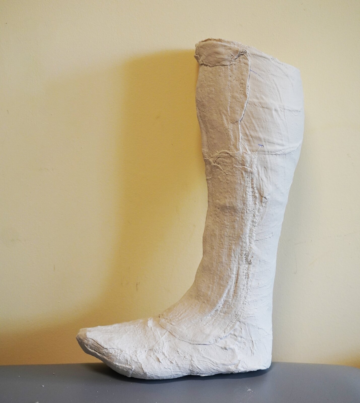 Zdjęcie przedstawia negatyw gipsowy lewej nogi - jej części poniżej kolana aż do stopy. Jest on wykonany z gipsu o białym zabarwieniu. Negatyw jest ustawiony pionowo na blacie.