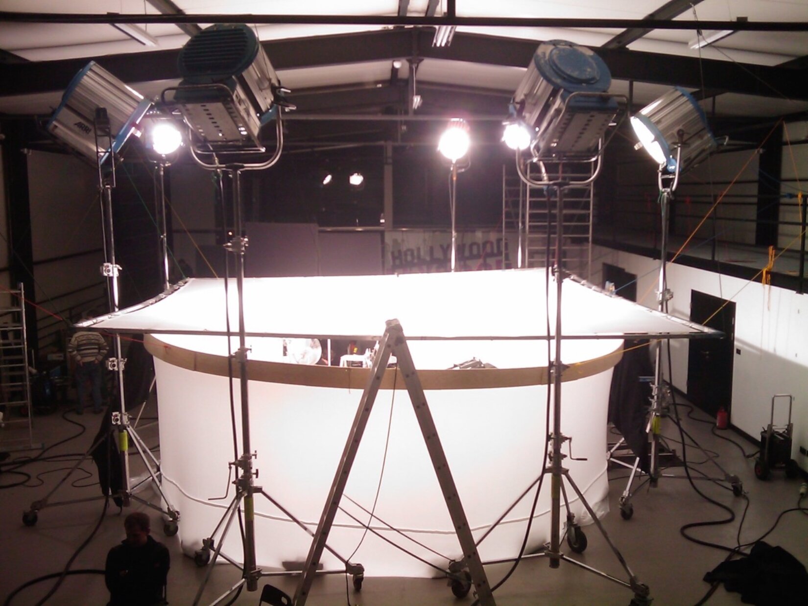 Ilustracja przedstawia oświetlenie spotu reklamowego w studio nagraniowym. Pomieszczenie posiada wysokie ściany i jest bardzo obszerne. Na środku pomieszczenia znajduje się oddzielone jasnym materiałem miejsce, w którym prowadzone są nagrywki. Jest ono oświetlone wysokimi lampami, które wydają bardzo mocne, białe i sztuczne światło. W studio widoczne są dwie osoby.