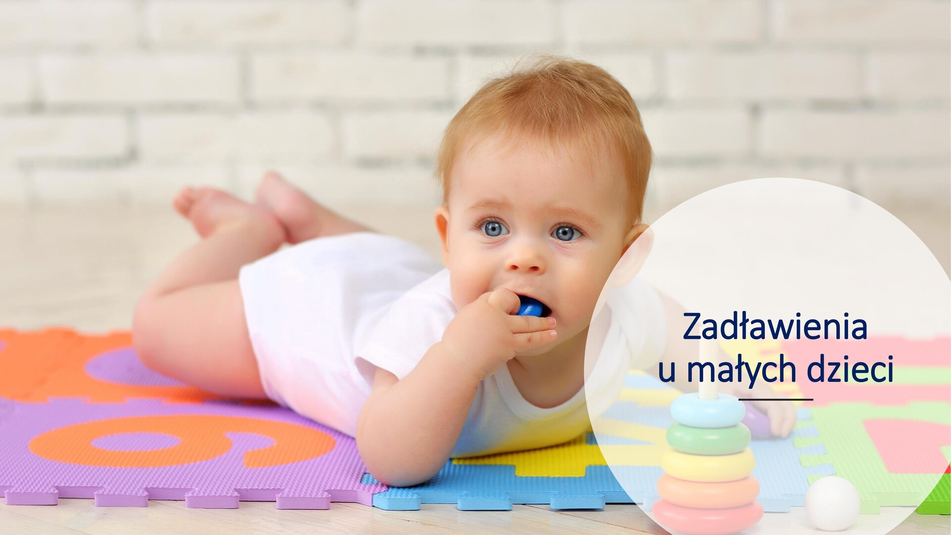 Slajd przedstawia zdjęcie małego chłopca leżącego na brzuchu. Jest rudy, a w ręku trzyma niebieską zabawkę, którą wkłada do buzi. Ubrany jest w białe śpioszki. Leży na kolorowej macie. W prawym dolnym rogu białe koło, a w nim napis: „Zadławienia u małych dzieci”.