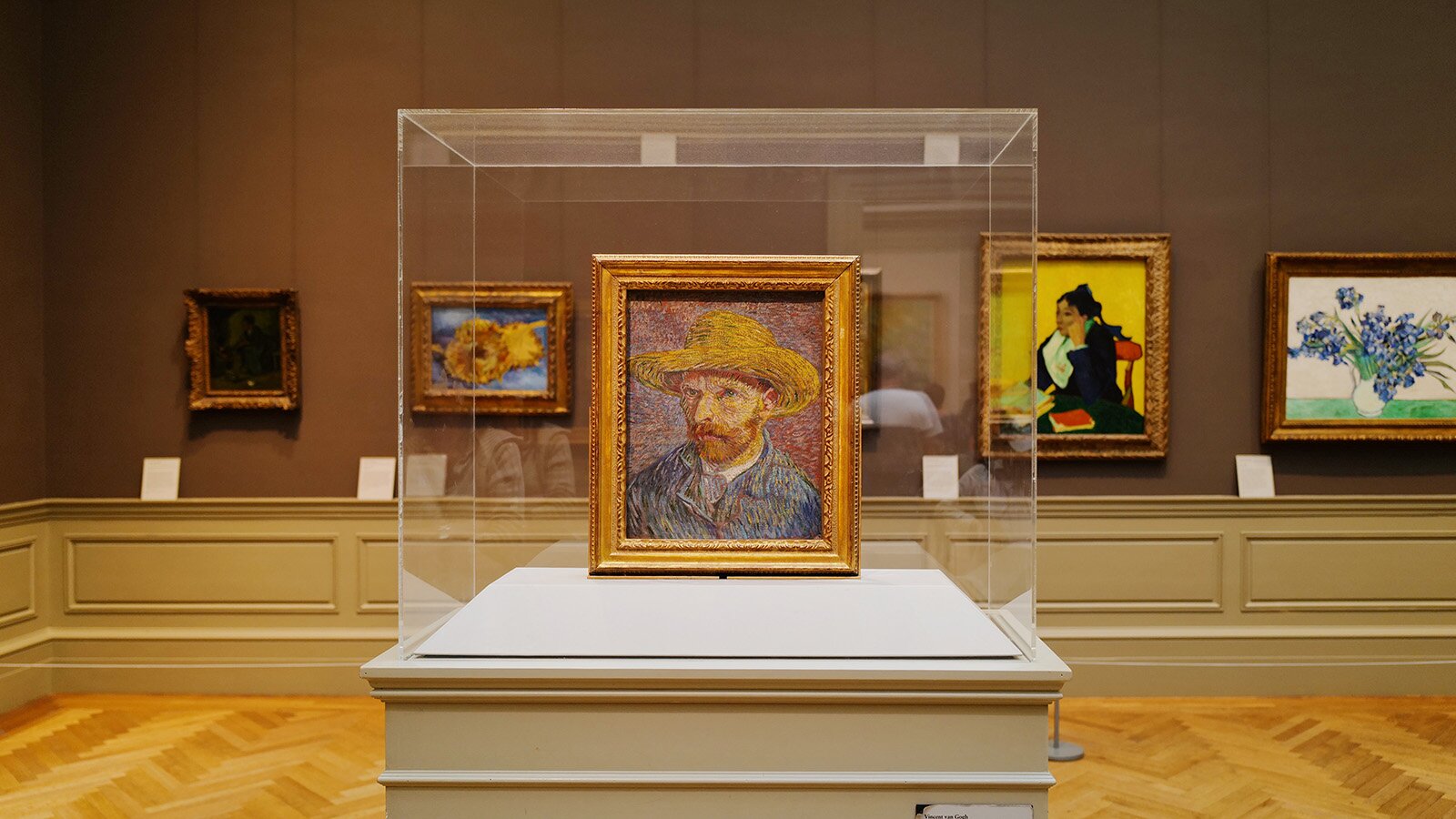 Zdjęcie przedstawia wnętrze galerii sztuki. Na ścianach wiszą obrazy w złotych ramach. Na środku w oszklonej gablocie widoczny jest obraz przedstawiający mężczyznę w kapeluszu. 