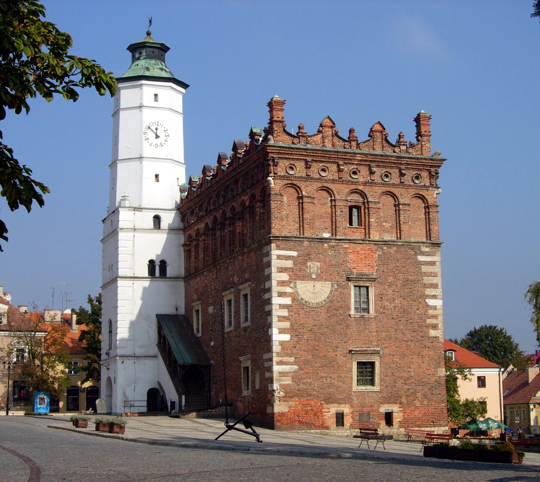 Na zdjęciu dwupiętrowy budynek z czerwonej cegły z dobudowaną wieżą w kolorze białym. U góry wieży zegar, dach w kolorze zielonym. Na jednej ze ścian budynku widoczny zegar słoneczny. Budynek zwieńczony attyką - zdobieniami osłaniającymi dach.