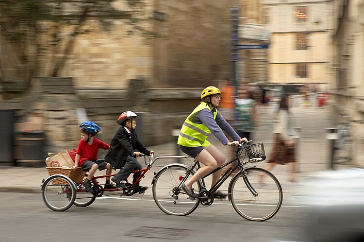 Zdjęcie przedstawia kobietę na rowerze ciągnącą przyczepkę z dziećmi, przypominającą rower bez przedniego koła. Kobieta w średnim wieku ma na sobie kask, kamizelkę odblaskową, ubrana jest ponadto w bluzkę na długi rękaw i krótkie, ciemne spodenki. Do jej roweru przyczepiony z przodu przyczepiony jest koszy, w tyłu natomiast jest pręt łączący się podwójnym rowerem dziecięcym. Kierownicę trzyma dziecko - chłopiec w kasku, ciemnej kurtce i długich ciemnych spodniach. Chłopiec siedzi na siodełku rowerowym, za nim siedzi kolejne dziecko - mniejszy chłopiec ubrany w kolorową bluzkę na krótki rękaw i krótkie spodenki. Chłopczyk wykręca głowę tak, że patrzy na to, co jest za nim. Prawą rękę opiera o wiklinowy kosz, który stanowi element roweru. Obaj chłopcy mają stopy na pedałach rowerowych. Tło zdjęcia jest zamazane, ale widać na nim jakieś budynki, chodnik i przechodzących ludzi. 