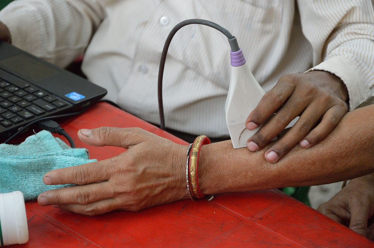 Zdjęcie przedstawia blat stołu przy którym siedzi dwoje ludzi: lekarz prowadzący badanie ultrasonograficzne i pacjent. Lekarz ma przed sobą otwarty laptop, w lewej ręce trzyma głowicę ultrasonografu. Dotyka nią lewej ręki i kości przedramienia pacjenta.