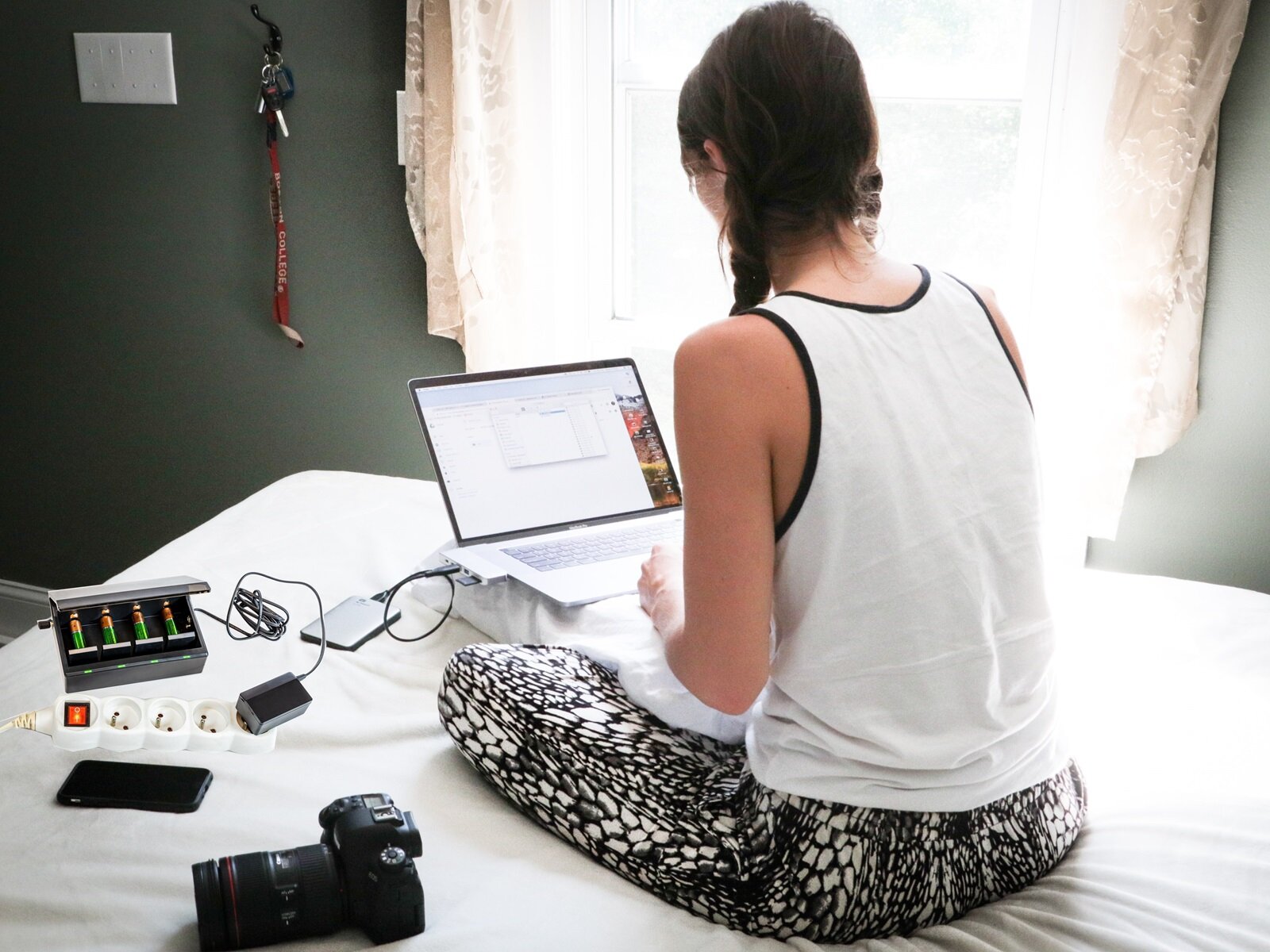 Ilustracja przedstawia siedząca na łóżku młodą kobietę, która pochyla się nad otwartym laptopem, który podłączony jest do zasilania przenośnego zwanego power bank. Widoczny jest również akumulator podłączony do gniazda elektrycznego ładujący baterie paluszki. Obok niego leży telefon komórkowy i aparat  fotograficzny. 