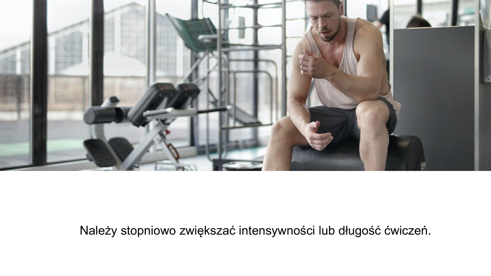 Biały slajd. W górnej części slajdu zdjęcie przedstawiające mężczyznę na siłowni. Siedzi on na ławeczce i ma zamknięte oczy. Prawą rękę opiera o udo, a lewy łokieć o kolano. W tle maszyny sportowe oraz przeszklona ściana, za którą widoczny jest skośny dach sąsiadującego budynku. W dolnej części slajdu tekst: „Należy stopniowo zwiększać intensywności lub długość ćwiczeń”.