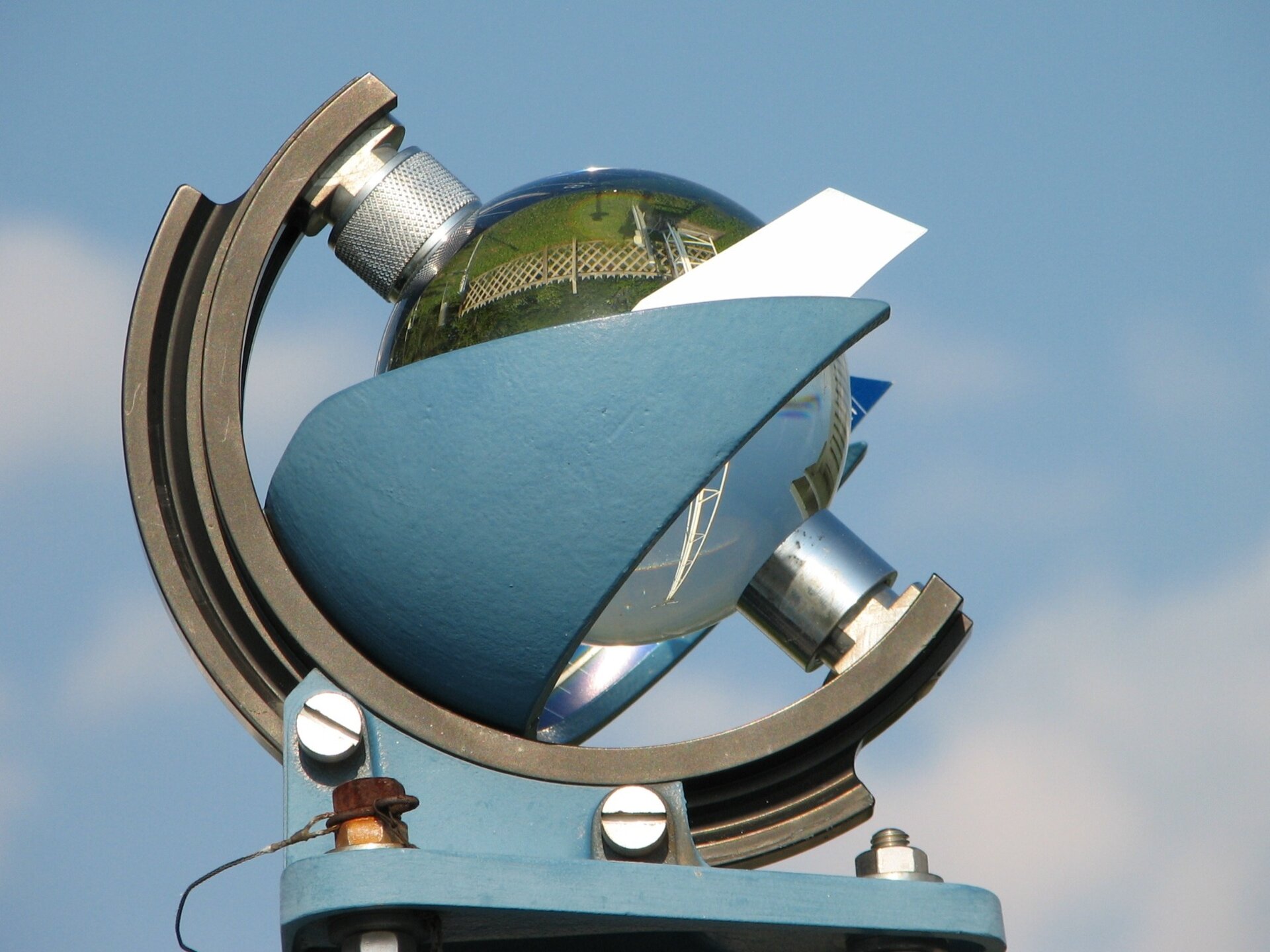 Fotografia przedstawiająca heliograf. Na kamienistym terenie stoi betonowa konstrukcja przedzielona ścianką. Po obu stronach stoją podobnie wyglądające urządzenia składające się z metalowej podstawy w kształcie litery T, metalowego ramienia w formie półksiężyca, na którym umieszczona jest szklana kula w taki sposób, by mogła się obracać jak globus. Kula z dwóch stron jest otoczona metalowymi, trójkątnymi osłonami.