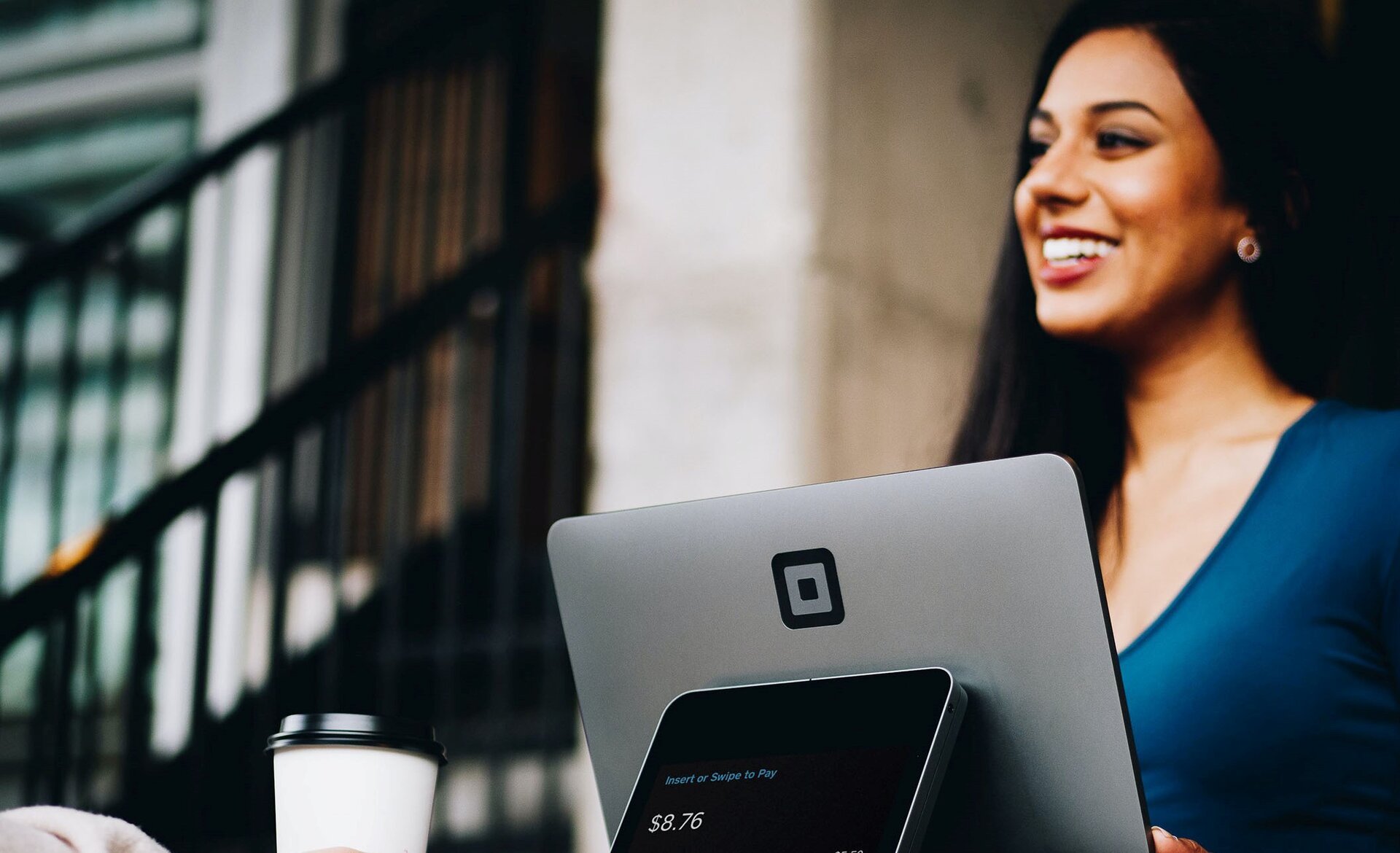 Zdjęcie przedstawia młodą kobietę, w ujęciu od klatki piersiowej w górę, zwróconą w lewo i stojącą za monitorem kasy. Kobieta ma długie, ciemne włosy, jest ubrana w niebieską bluzkę i szeroko się uśmiecha. Przed nią widoczny jest monitor kasy. O tył monitora oparty jest drugi, mniejszy ekran, na którym widoczny jest napis w języku angielskim: Insert or Swipe to Pay oraz kwota. Na lewo od dwóch monitorów widoczny jest jednorazowy kubek.