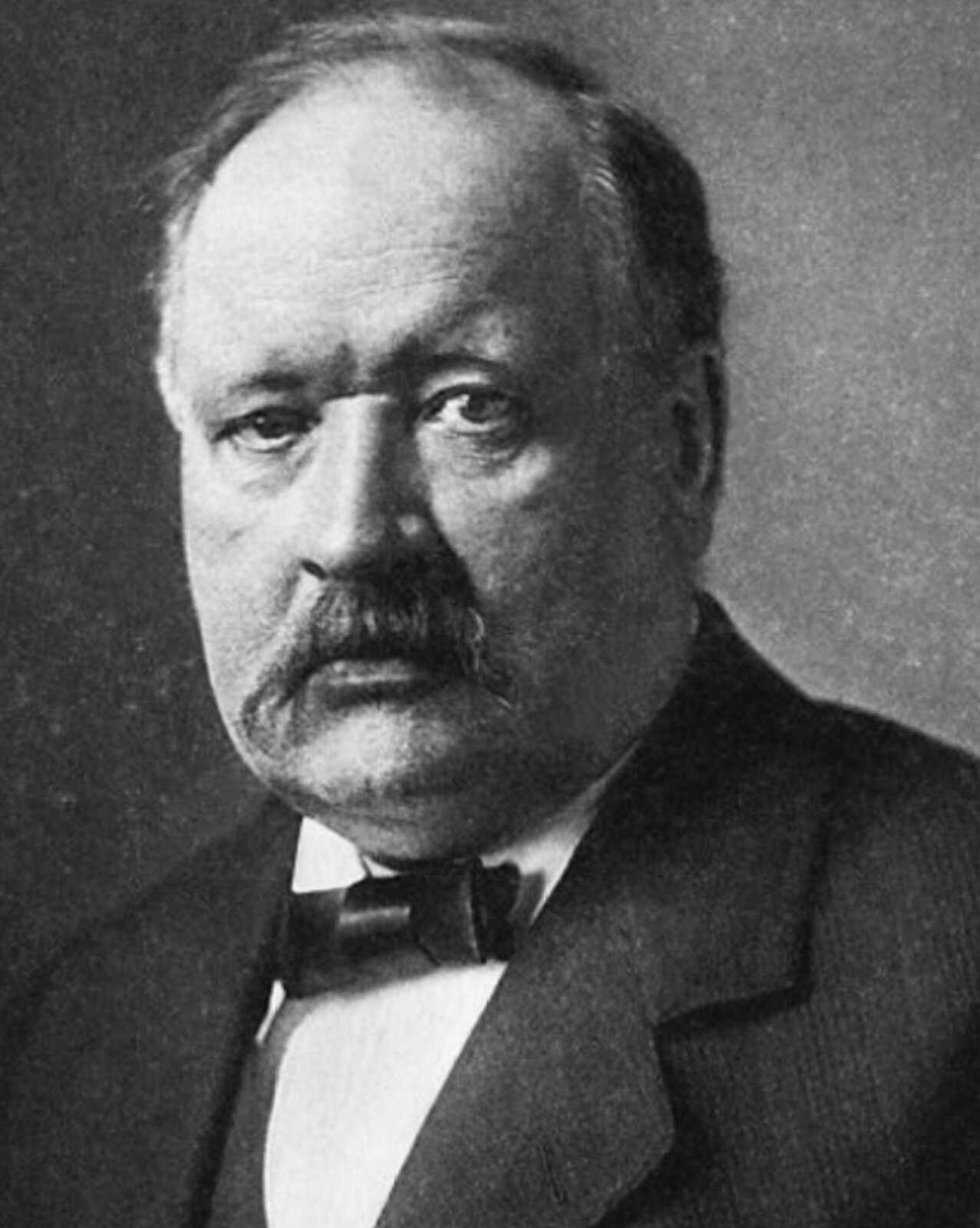 Fotografia czarno-biała przedstawiająca Svante'a Augusta Arrheniusa. Mężczyzna w średnim wieku, lekko wyłysiały z krótkimi włosami oraz wąsami. Ma lekko zmarszczone czoło i zaokrągloną twarz. Ubrany jest w białą koszulę, czarny garnitur oraz muchę.