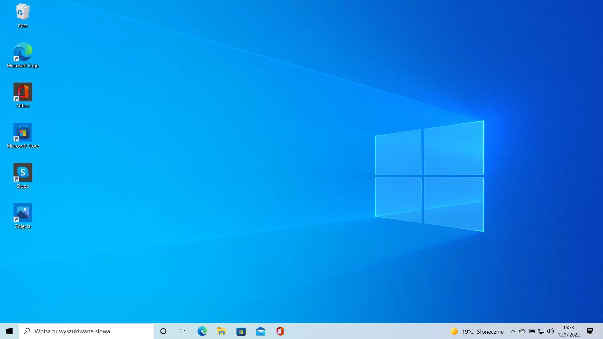 Na zdjęciu przedstawiono zrzut pulpitu dla systemu Windows 10. Na zdjęciu widać niebieską tapetę.  Na tapecie po prawej stronie widocznie jest białe logo Windows. Na tapecie, w lewym górnym rogu znajdują się ikony podpisane jako: kosz, Microsoft Edge, Office, Microsoft Store Skype, zdjęcia.Poniżej tapety znajduje się pasek narzędzi. Po lewej stronie paska znajduje się ikony: czarne logo Windows, pole wyszukiwania z napisem wpisz tu wszystkie słowa, czarny okrąg, trzy pionowe prostokąty z prostą po prawej stronie, okrąg przypominający zawijająsą się fale, żółty folder, teczka z kolorowym logiem Windows, koperta, pomarańczowy prostopadłościan bez ściany przedniej i tylnej który jest widoczny pod kątem. Po prawej stronie widoczne są ikony: słońce, napis: dziewiętnaście stopni Celsjusza słonecznie, trójkąt po rozwinięciu którego pojawiają się dodatkowe opcje, chmura, bateria z podłączonym kablem, ekran z kablem LAN, głośnik, godzina i data przykładowo piętnasta trzydzieści trzy, dwutysięczny dwudziesty drugi lipiec dwunasty, czarna karka z białym napisem i księżycem w prawym dolnym rogu.