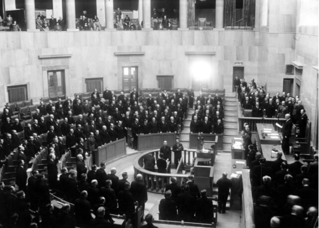 Zdjęcie przedstawia salę parlamentarną sejmu polskiego. Ławy poselskie rozmieszczone są na sali w kształcie półkola. W centralnej części półkola znajduje się mównica. Nad nią, wyżej znajduje się stół, za którym zasiada marszałek sejmu. Wszyscy posłowie na zdjęciu stoją, marszałek odczytuje dokument. Na balkonie sali zasiadają oficerowie i zgromadzeni goście. Sala jest wysoka, na balkonach widoczne są kolumny. 