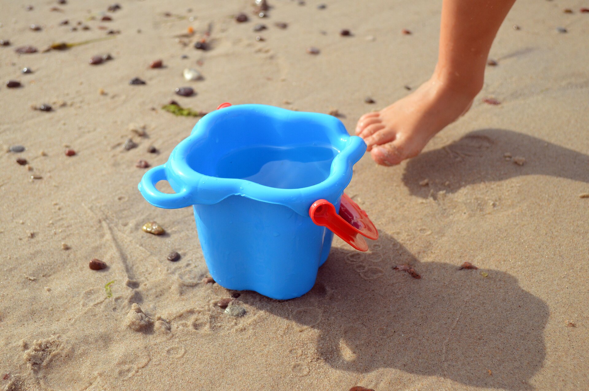 Rys. 6. Zdjęcie poglądowe przedstawia fragment plaży z piaskiem i stopę dziecka obok wiaderka napełnionego wodą.