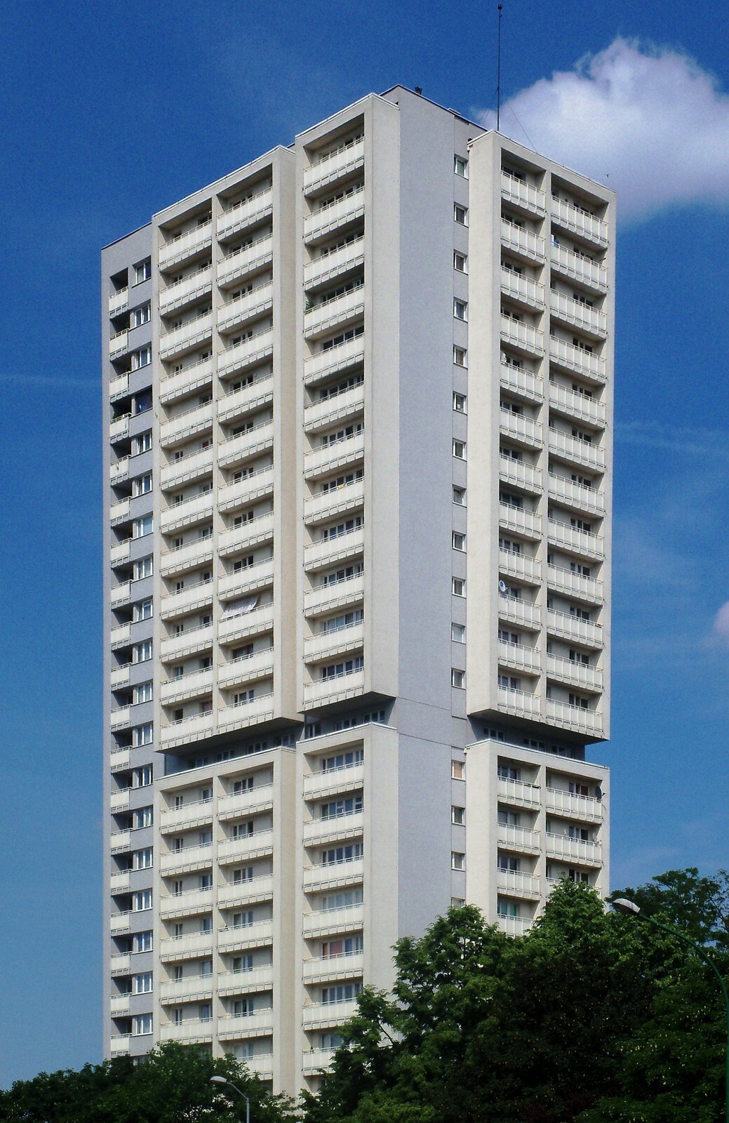 Wieżowiec w Katowicach to wysoki budynek z oknami i balkonami ustawionymi bardzo blisko siebie. Z jednej strony są to rzędy po cztery balkony, z drugiej - po dwa balkony z rzędem okien. Budynek jest szaro - beżowy, z białymi balkonami. Na dole rosną drzewa.