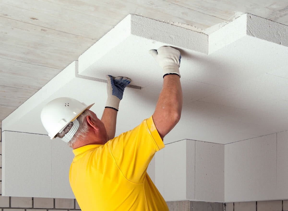 Zdjęcie przedstawia izolacyjny beton komórkowy. Widać pracownika, który płytę betonu nakłada na sufit. Pracownik ma na głowie kask.