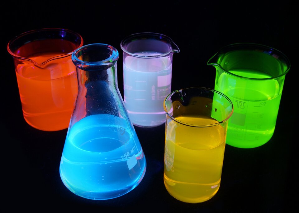 Na zdjęciu znajdują się wodne roztwory związków o zdolności do fluorescencji w świetle UV. Roztwory znajdują się w walcowatych zlewkach oraz w kolbie stożkowej w postaci stożka zakończonego wąskim kominem. Mają one kolor czerwony, niebieski, biały, żółty oraz zielony. Tło zdjęcia jest czarne.