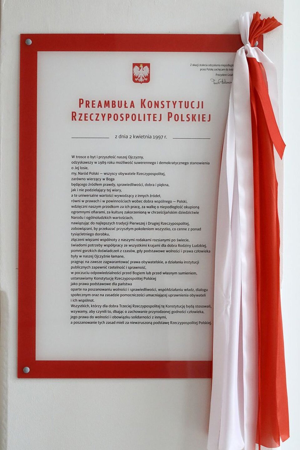 Zdjęcie przedstawia ścianę, na której umieszczona jest tablica z tekstem Preambuły Konstytucji Rzeczypospolitej Polskiej. Tekst obramowany jest czerwoną wstążką. Po prawej stronie widnieje biało‑czerwona szarfa. Tekst na ilustracji jest czytelny. Jego zapis brzmi: 
Preambuła Konstytucji Rzeczypospolitej Polskiej
z dnia 2 kwietnia 1997 r. 
W trosce o byt i przyszłość naszej Ojczyzny,
odzyskawszy w 1989 roku możliwość suwerennego i demokratycznego stanowienia o Jej losie,
my, Naród Polski - wszyscy obywatele Rzeczypospolitej,
zarówno wierzący w Boga będącego źródłem prawdy, sprawiedliwości, dobra i piękna,
jak i nie podzielający tej wiary, a te uniwersalne wartości wywodzący z innych źródeł,
równi w prawach i w powinnościach wobec dobra wspólnego – Polski,
wdzięczni naszym przodkom za ich pracę, za walkę o niepodległość okupioną ogromnymi ofiarami, za kulturę zakorzenioną w chrześcijańskim dziedzictwie Narodu i ogólnoludzkich wartościach,
nawiązując do najlepszych tradycji Pierwszej i Drugiej Rzeczypospolitej,
zobowiązani, by przekazać przyszłym pokoleniom wszystko,
co cenne z ponad tysiącletniego dorobku,
złączeni więzami wspólnoty z naszymi rodakami rozsianymi po świecie, świadomi potrzeby współpracy ze wszystkimi krajami dla dobra Rodziny Ludzkiej,
pomni gorzkich doświadczeń z czasów, gdy podstawowe wolności i prawa człowieka były w naszej Ojczyźnie łamane,
pragnąc na zawsze zagwarantować prawa obywatelskie,
a działaniu instytucji publicznych zapewnić rzetelność i sprawność,
w poczuciu odpowiedzialności przed Bogiem lub przed własnym sumieniem,
ustanawiamy Konstytucję Rzeczypospolitej Polskiej jako prawa podstawowe dla państwa oparte na poszanowaniu wolności i sprawiedliwości,
współdziałaniu władz, dialogu społecznym oraz na zasadzie pomocniczości umacniającej uprawnienia obywateli i ich wspólnot.
Wszystkich, którzy dla dobra Trzeciej Rzeczypospolitej tę Konstytucję będą stosowali,
wzywamy, aby czynili to, dbając o zachowanie przyrodzonej godności człowieka, jego prawa do wolności i obowiązku solidarności z innymi,
a poszanowanie tych zasad mieli za niewzruszoną podstawę Rzeczypospolitej Polskiej.