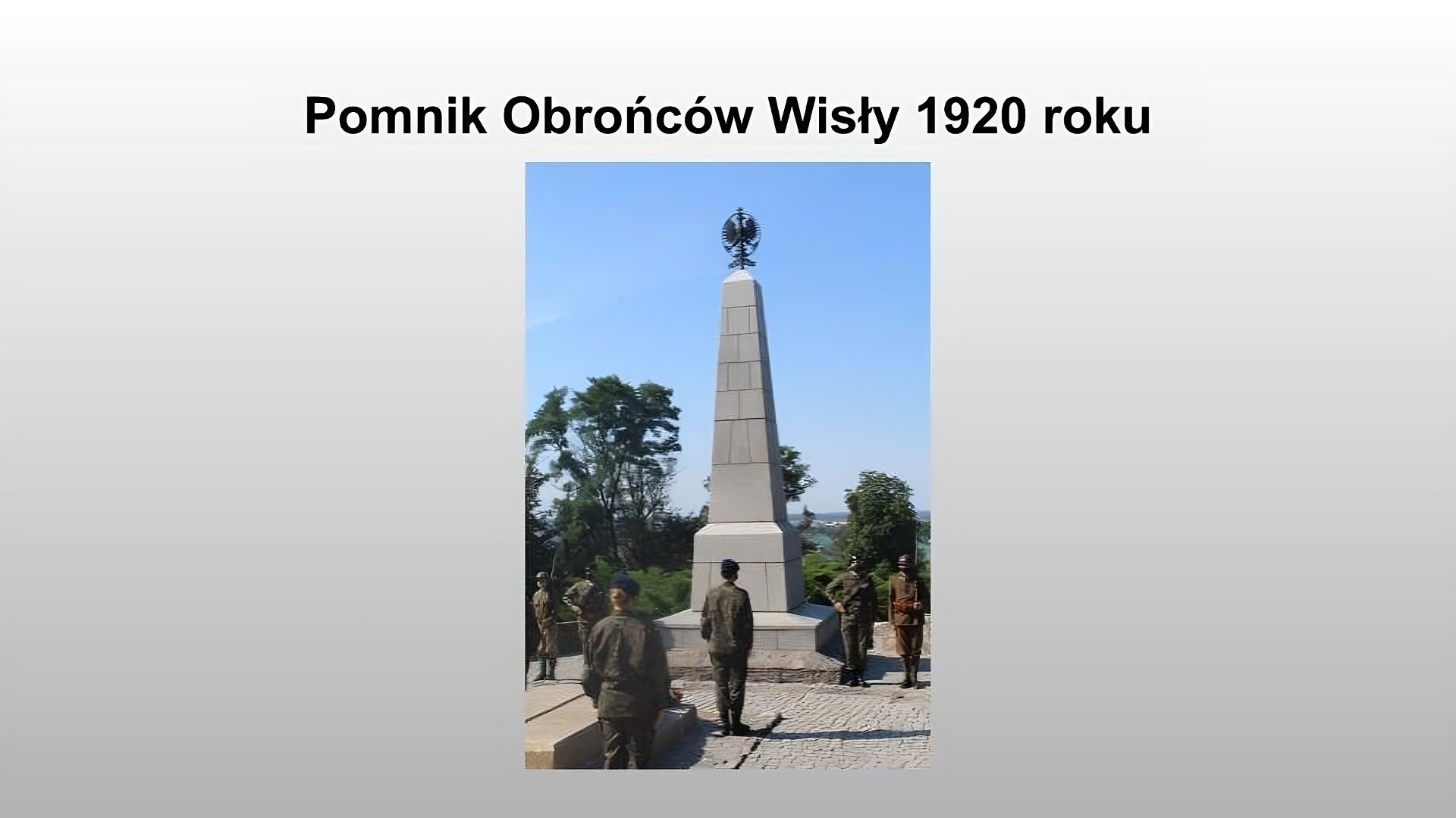 Jasnoszary slajd. Na górze slajdu nagłówek: „Pomnik Obrońców Wisły 1920 roku”. Poniżej zdjęcie przedstawiające szary, wysoki pomnik przypominający obelisk. Na jego szczycie metalowa rzeźba w kształcie godła polskiego. Po przed i bokach pomnika stoją żołnierze w mundurach. W tle drzewa. 