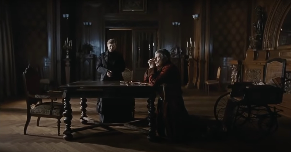 Fotografia przedstawia kadr z filmu, w którym ukazanych jest dwóch mężczyzn pogrążonych w zadumie. Starszy z nich klęczy przy stole, wspierając się łokciami o jego blat. Ujęty bokiem do widza, ubrany jest w ciemnobordowy szlafrok, który nadaje mu domowy, lecz dostojny wygląd. Naprzeciw niego, po drugiej stronie stołu, stoi krzesło z wysokim, rzeźbionym oparciem – puste, jakby symbolicznie czekające na kogoś.

Po prawej stronie klęczącego mężczyzny stoi drugi – młodszy, ze złożonymi rękami i głową spuszczoną w dół. Jego ciemny surdut z szerokim kołnierzem podkreśla powagę sytuacji. Obaj znajdują się w dużym, przyciemnionym salonie, którego atmosfera budowana jest przez detale: ozdobne kinkiety na ścianach, wysokie świeczniki ustawione wzdłuż ścian oraz eleganckie, klasyczne meble.