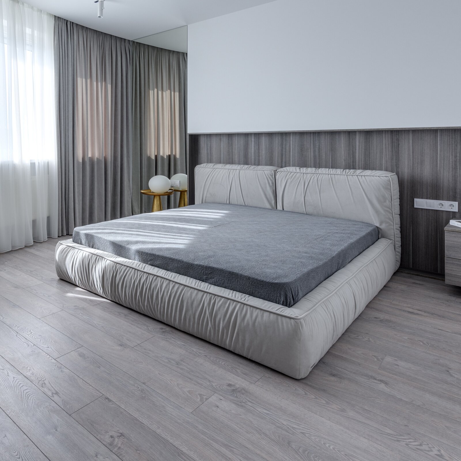 Na ilustracji znajduje się sypialnia. Pośrodku stoi duże łóżko. Koło łóżka stoi mała szafka, a z drugiej strony mały stolik. W dużym oknie wiszą białe firanki  i szare zasłony.