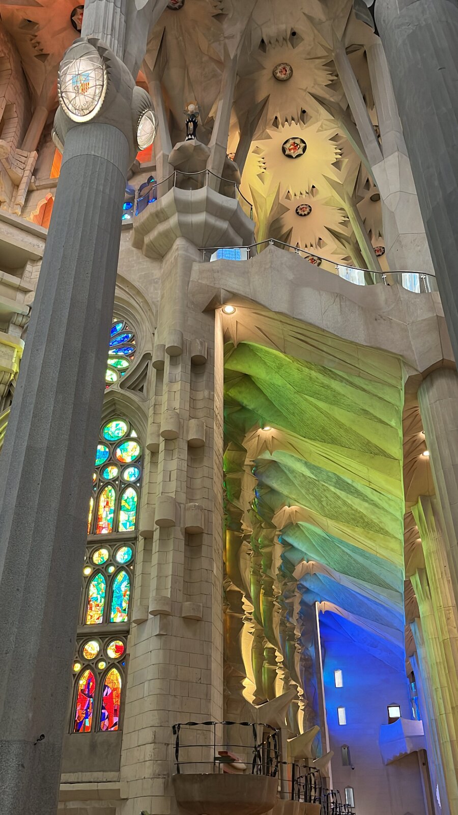 Ilustracja przedstawia wnętrze katedry Sagrada Família. Ukazuje wysokie kolumny przypominające pnie drzew, które unoszą złożone sklepienie o geometrycznych wzorach. Kolorowe witraże w odcieniach czerwieni, zieleni, błękitu i żółci przepuszczają światło, tworząc na kamiennych powierzchniach intensywne, wielobarwne refleksy. Gra światła podkreśla rytm kolumn i wysokość przestrzeni, nadając wnętrzu wrażenie ruchu i lekkości.