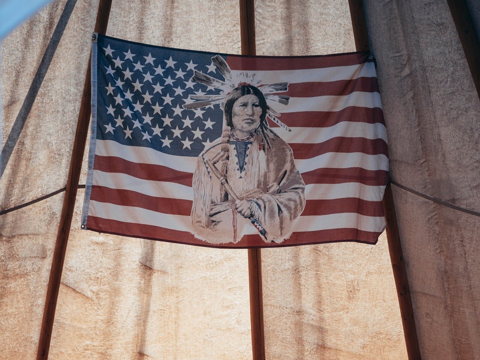 Zdjęcie przedstawia amerykańską flagę zawieszoną na oświetlonym od tyłu materiale, prawdopodobnie części namiotu. Pośrodku flagi widoczna jest postać rdzennego mieszkańca Ameryki, w ujęciu od pasa w górę, który przedstawiony jest w tradycyjnym stroju, w pióropuszu na głowie i z tomahawkiem w lewej dłoni.