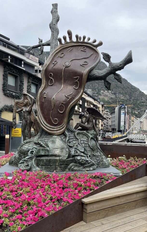 Ilustracja przedstawia fragment placu, na którym znajduje się pomnik. Pomnik przedstawia tarczę zegara zawieszoną na konarach drzewa. Tarcza jest ogromnych rozmiarów i mimo, że wykonana z metalu jej forma zawieszona na drzewie sprawia wrażenie, że jest z miękkiej tkaniny, ponieważ jest pofalowana. Na planie pierwszym znajduje się klomb z kwiatami, a w oddali ulica z zabudowaniami. Tło stanowią góry oraz jasne niebo.