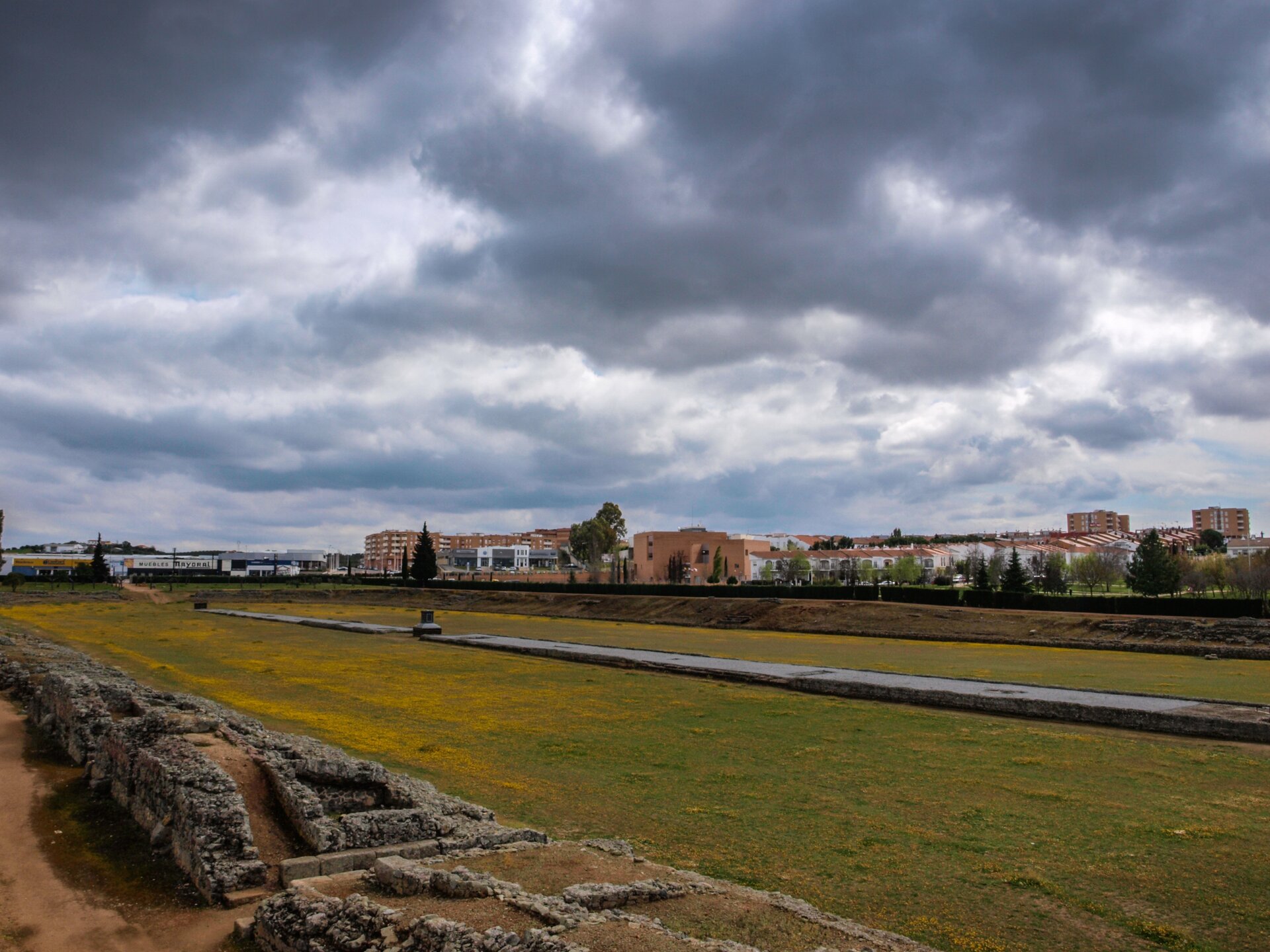 Fotografia przedstawia pozostałości po cyrku rzymskim, który ma kształt długiego prostokąta. Wewnątrz toru biegnie kamienny pas, który dzieli go na dwie części. Za torem – pozostałości po trybunach. W tle współczesne zabudowania miejskie. Fotografia została wykonana w pochmurny dzień.