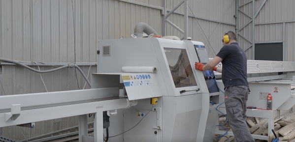 Zdjęcie przedstawia obrabiarkę. Jest to duże urządzenie, przez które przechodzi drewno. Przy ekranie urządzenia stoi pracownik w słuchawkach ochronnych.