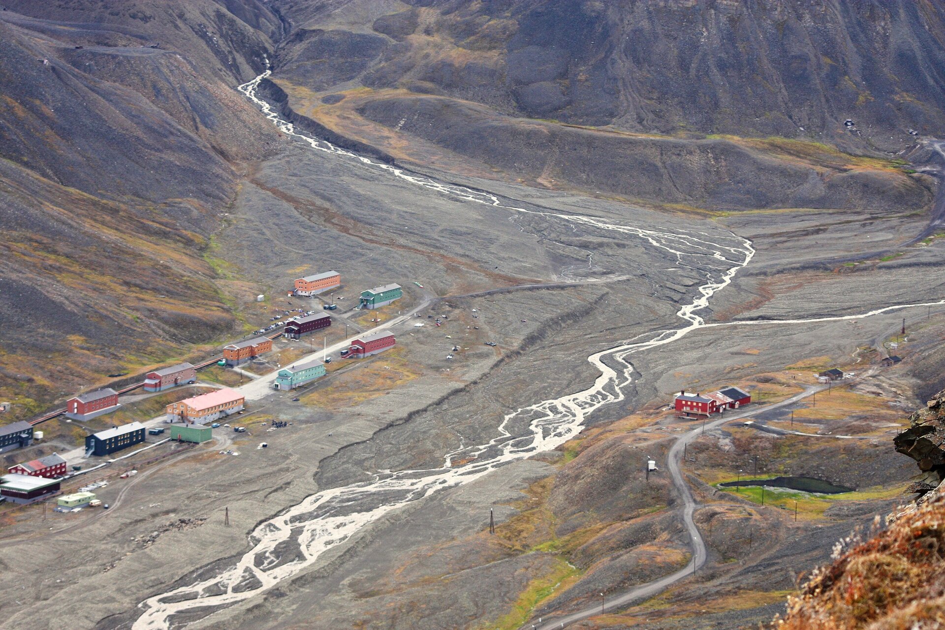 Zdjęcie przedstawia dolinę rzeki warkoczowej Longyear (Spitsbergen), zasilana wodami lodowców Longyearbreen i Larsbreen. Na zdjęciu skaliste góry, między którymi znajduje się dolina. Doliną płynie niewielki strumień wody. Obok niego niewielkie zabudowania mieszkalne. 