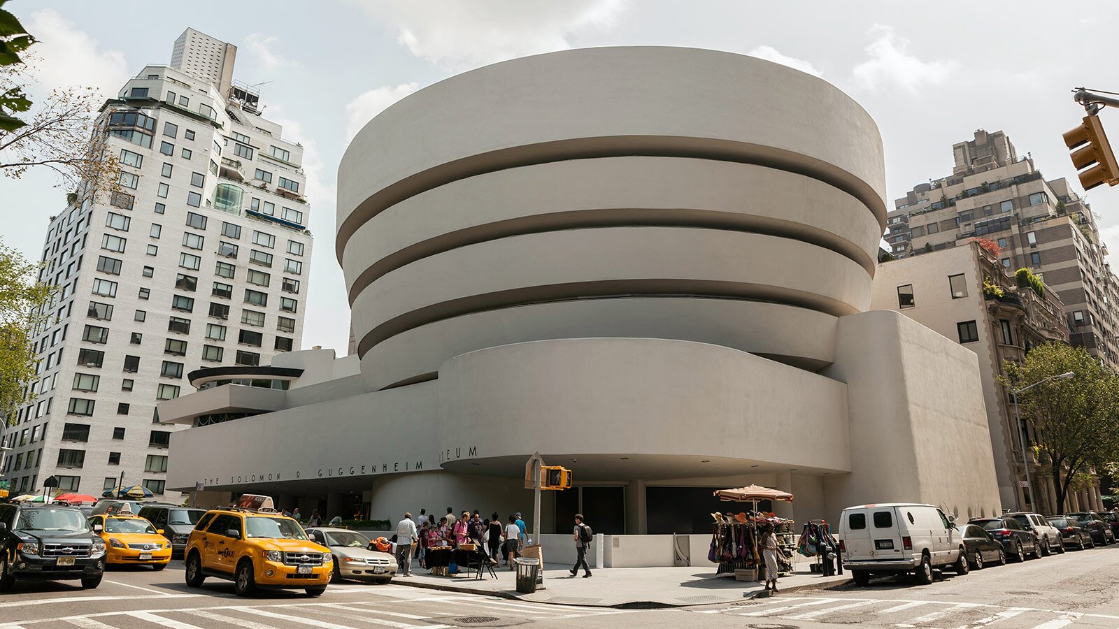 Zdjęcie przedstawia narożny budynek w formie rotundy, osadzonej na rozłożystej, wielosegmentowej podstawie. Rotunda przypomina cylindrycznie zwiniętą wstęgę, rozszerzającą się ku górze. W dolnej części budynku, po lewej stronie, widoczne jest wejście, nad którym na elewacji znajduje się napis w języku angielskim: THE SOLOMON R. GUGGENHEIM MUSEUM. Po obu jego stronach widoczne są wielopiętrowe budynki. Na krzyżujących się ulicach poruszają się samochody, a po chodnikach spacerują ludzie.