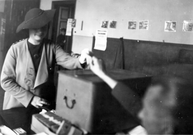 Zdjęcie przedstawia wnętrze sali. Kobieta w kapeluszu podaje osobie siedzącej za pulpitem kartę.