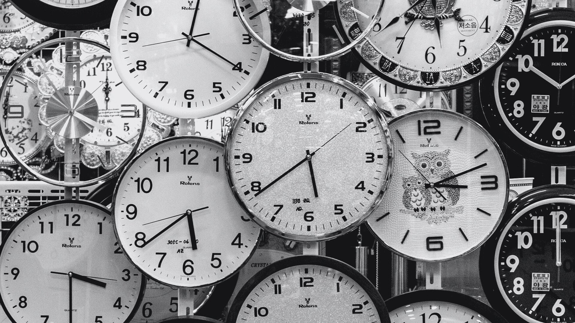 Czarno‑białe zdjęcie przedstawia kilkanaście tarcz zegarów ściennych zawieszonych jeden na drugim. Niektóre tarcze zachodzą na siebie. Wskazówki zegarów wskazują różne godziny. 