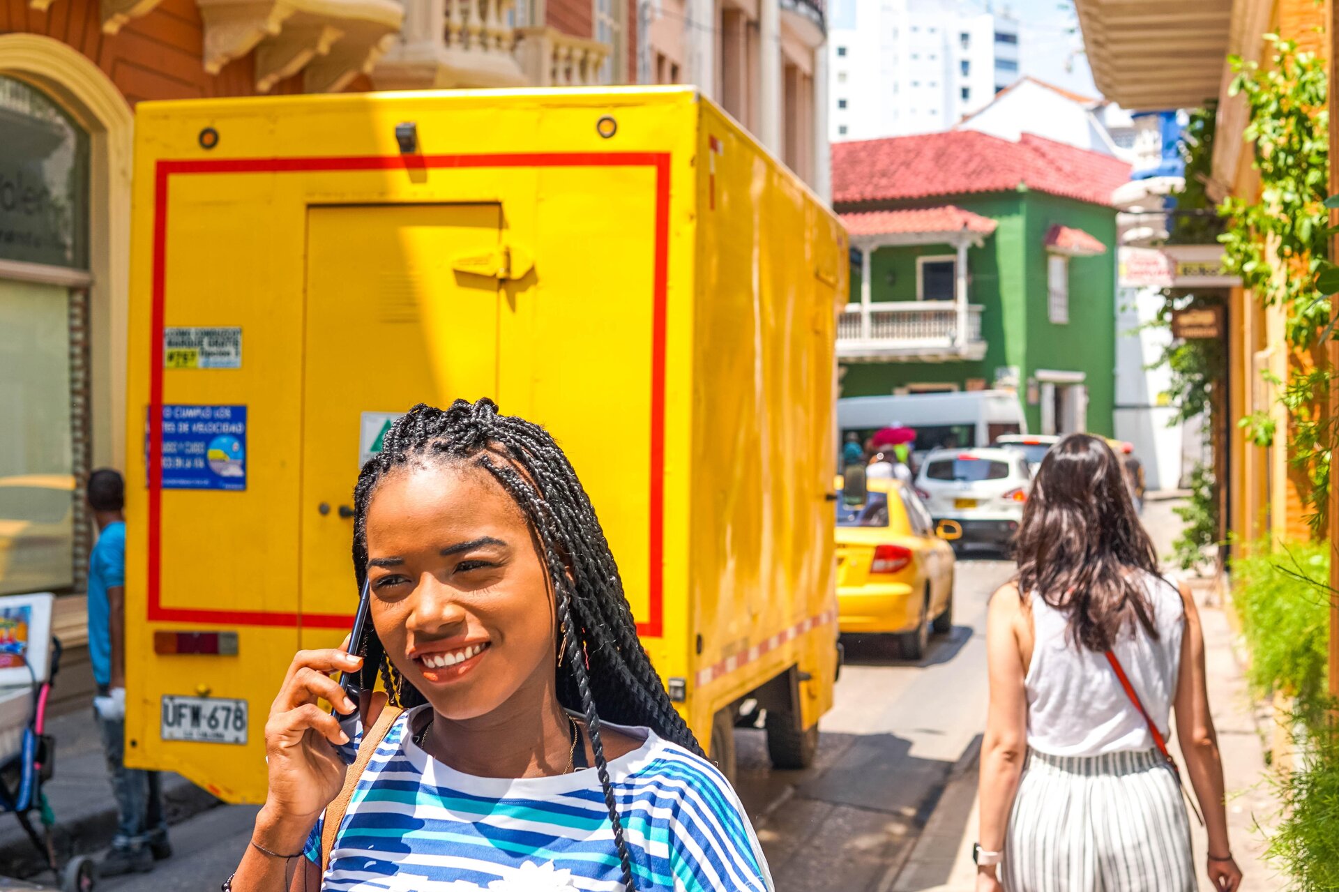 Zdjęcie przedstawia nastoletnią dziewczynę, w ujęciu od klatki piersiowej w górę, idącą ulicą i trzymającą telefon przy prawym uchu. Dziewczyna ma długie, ciemne włosy zaplecione w warkoczyki, jest ubrana w bluzkę w białe, niebieskie i ciemnoniebieskie paski, uśmiecha się. W tle za nią jest widoczny fragment ulicy oraz chodnika. Na ulicy są widoczne samochody dostawcze i osobowe. Na chodniku widoczna jest postać kobiety, w ujęciu od tyłu. Po obu stronach ulicy widoczne są kolorowe budynki.