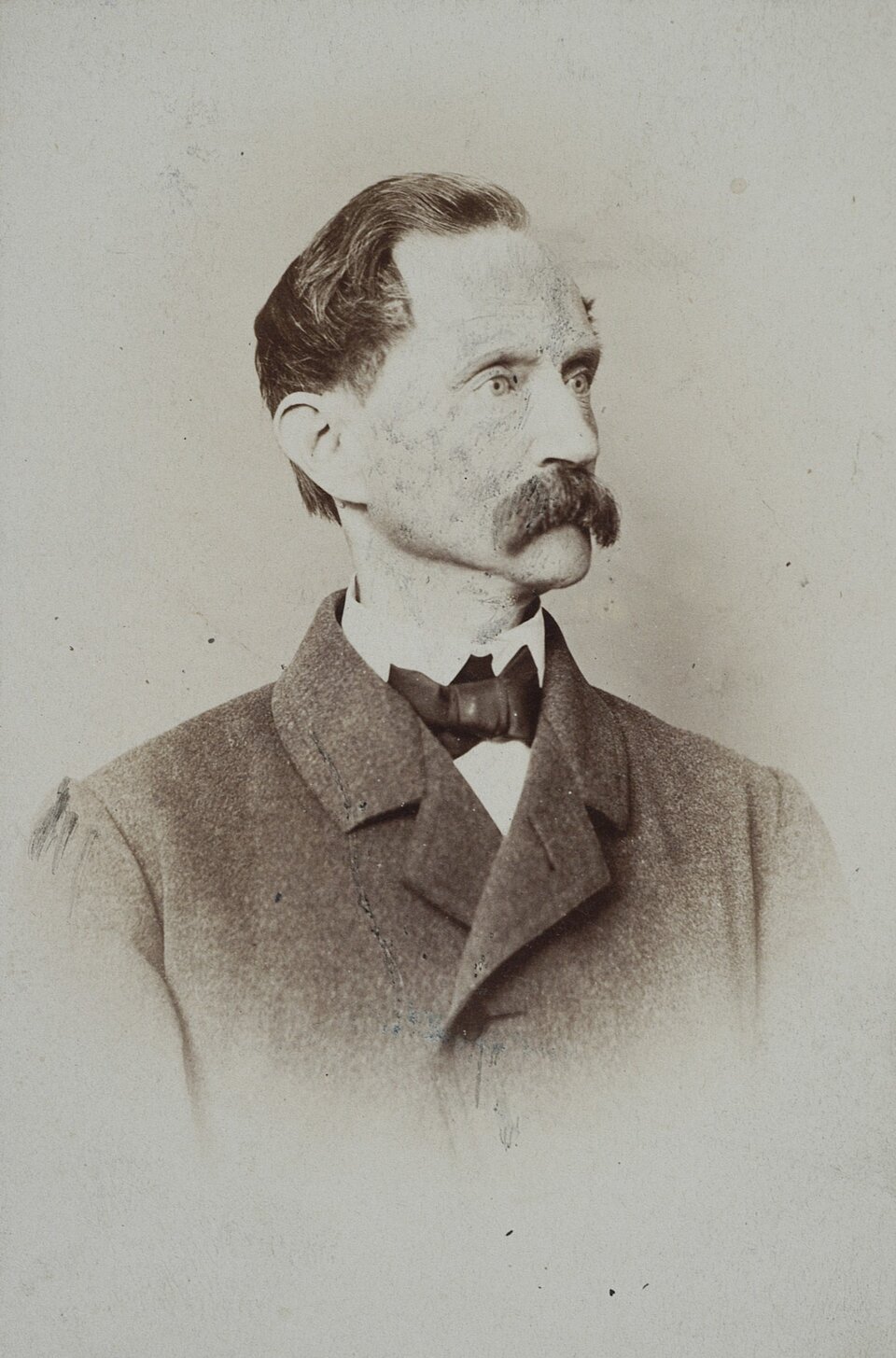 Zdjęcie przedstawia popiersie starszego mężczyzny z twarzą zwróconą w prawą stronę. Mężczyzna ma dłuższe włosy zaczesane do tyłu oraz sumiaste wąsy. Ubrany jest  w białą koszule z muszką oraz szara marynarkę. 