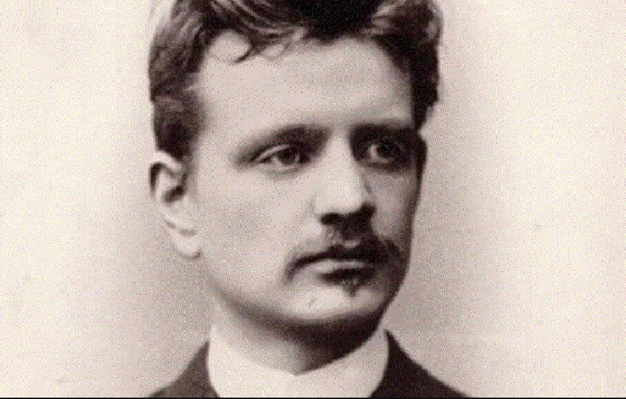 Fotografia przedstawia Jana Sibeliusa - ujęcie głowy i szyi. Młody mężczyzna o szczupłej twarzy, bujnych włosach, ma wąsy i drobny zarost na brodzie.