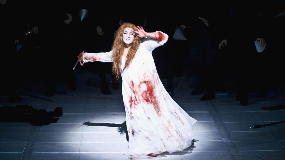 Ilustracja przedstawia scenę obłędu z opery G. Donizettiego Łucja z Lammermoore. Kobieta w białej, długiej, okrwawionej sukni. Stoi ma w ręce nóż, drugą ręką zasłania oczy przed światłem. Kobieta ma długie, rude włosy.