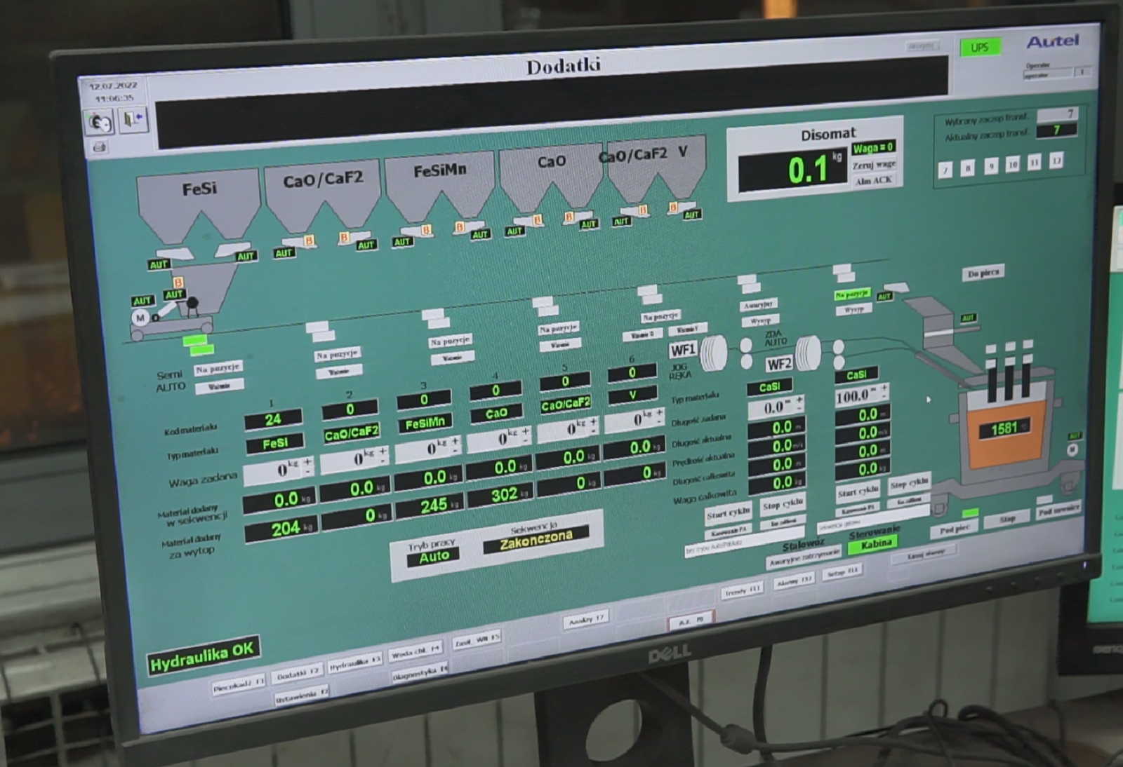 Na zdjęciu widać ekran z ilością wysypywanego materiału. Na ekranie widać okno z dedykowanego oprogramowania podpisane u góry: dodatki. Niżej znajdują się rysunki dwukomorowych magazynów z nazwami chemicznymi dodatków. Poniżej znajdują się ekrany na których wyświetlane są wartości liczbowe nastaw. Na dole olana znajduje się pasek narzędzi z dodatkowymi przyciskami.