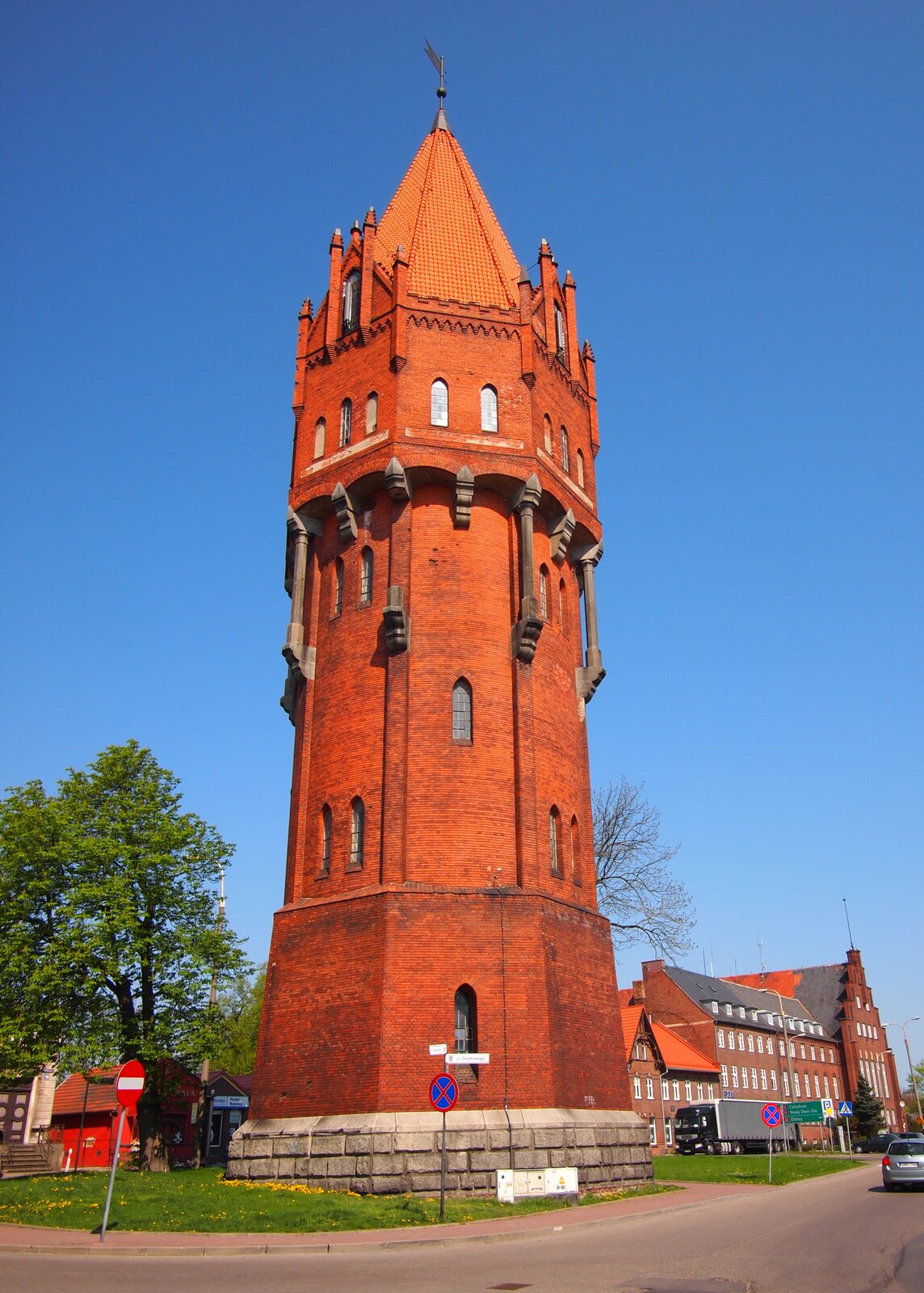 Zdjęcie przedstawia wieżę ciśnień. Wieża zajmuje całą wysokość zdjęcia. Zbudowana z czerwonej cegły. Ozdobnie wykończona. W tle błękitne niebo. Z lewej strony wieży zielone drzewo liściaste Z prawej strony, w tle, budynek z czerwonej cegły. Tuż przed wieżą ulica.