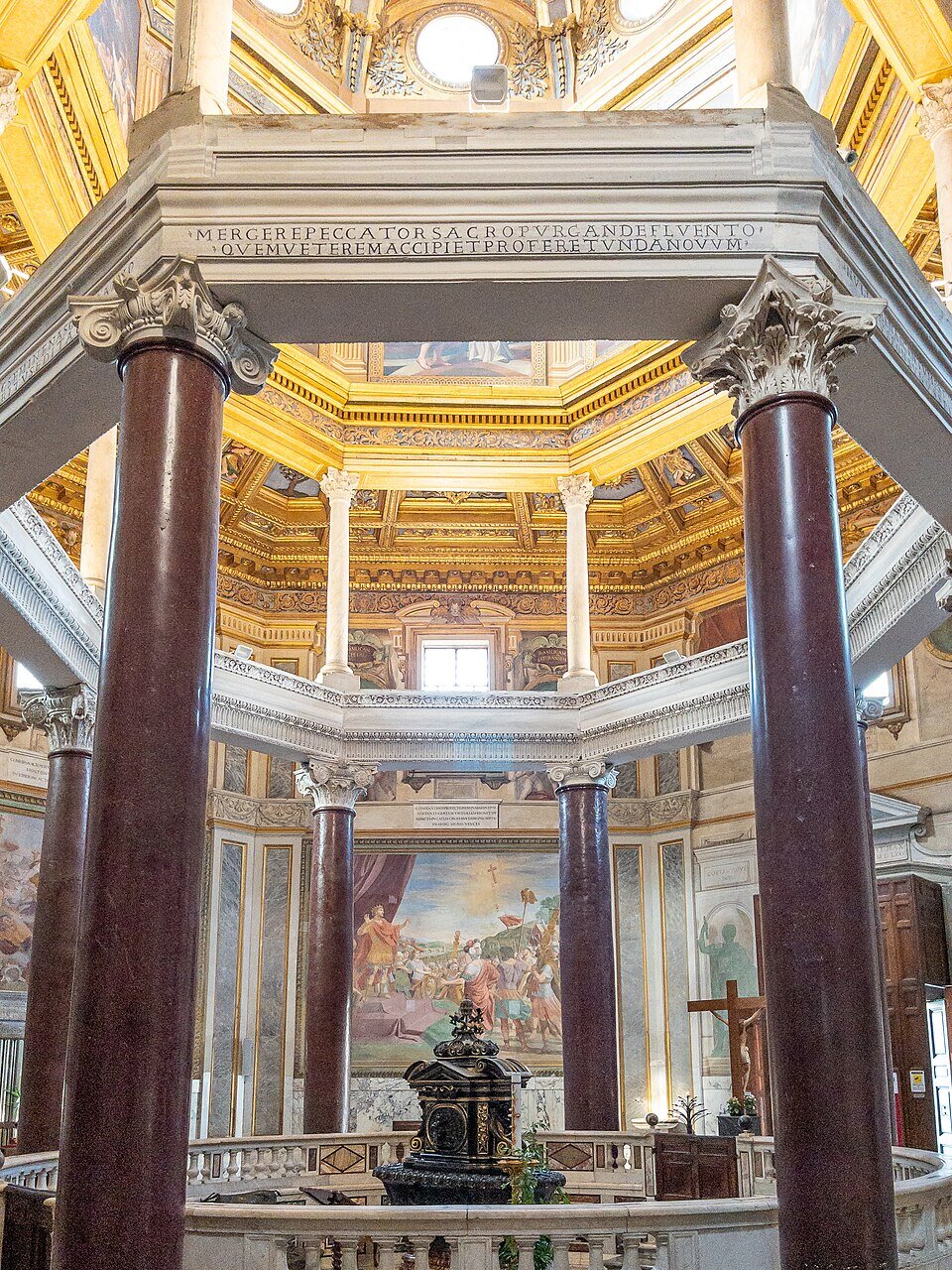 Bogato zdobione wnętrze baptysterium o planie centralnym: masywne, czerwone kolumny z korynckimi kapitelami podtrzymują architraw i górny pierścień arkad. W centrum znajduje się kamienna chrzcielnica otoczona balustradą, a ściany i sufit pokrywają złocenia, freski oraz dekoracje architektoniczne. Światło wpada przez okrągłe okna pod kopułą, podkreślając symetrię i monumentalny charakter przestrzeni.