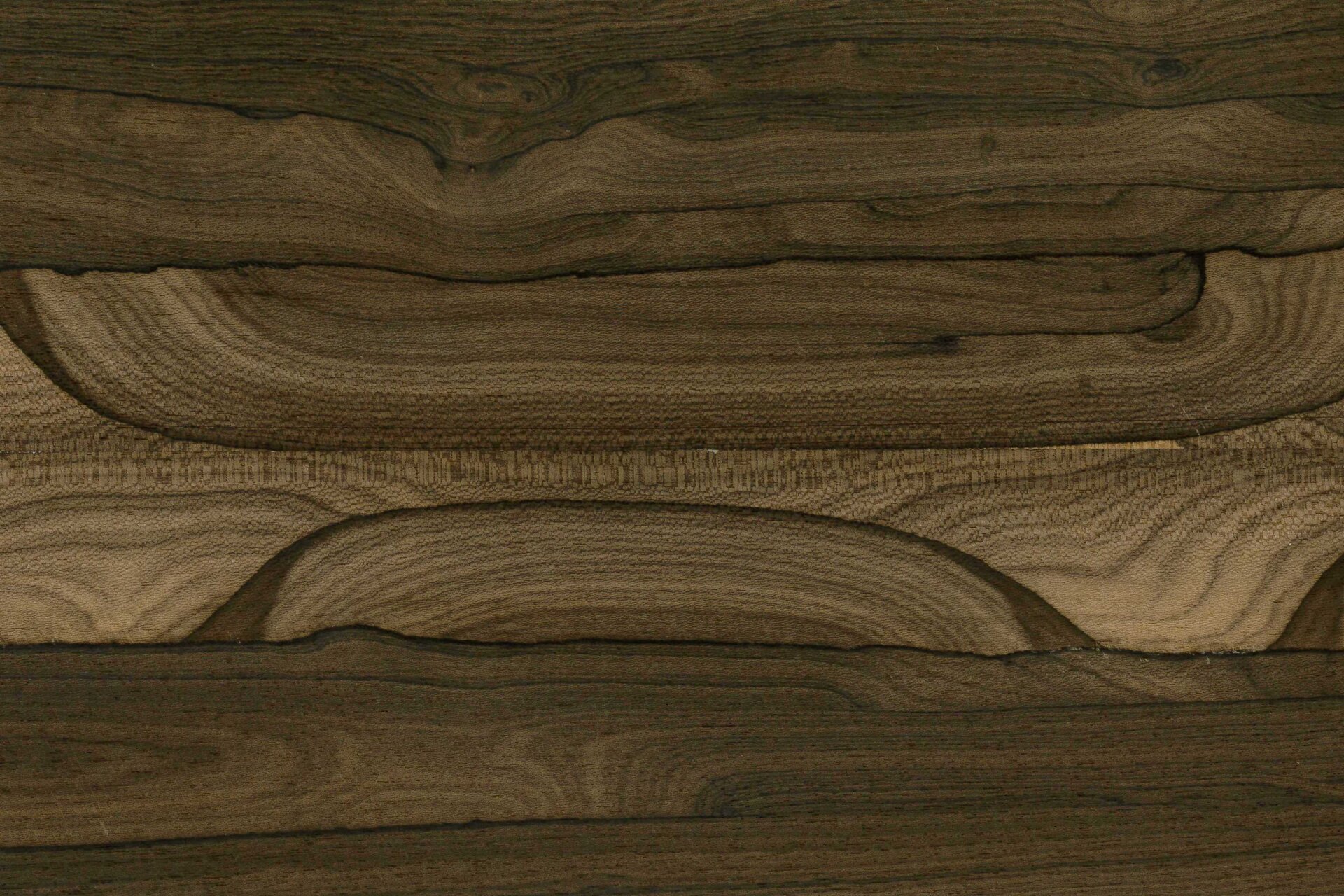 Zdjęcie przedstawia zbliżenie na okleinę imitującą drewno. Jest to cienki arkusz forniru. Okleina charakteryzuje się wzorem i teksturą przypominającą prawdziwe drewno. Posiada ona na swojej powierzchni między innymi słoje.