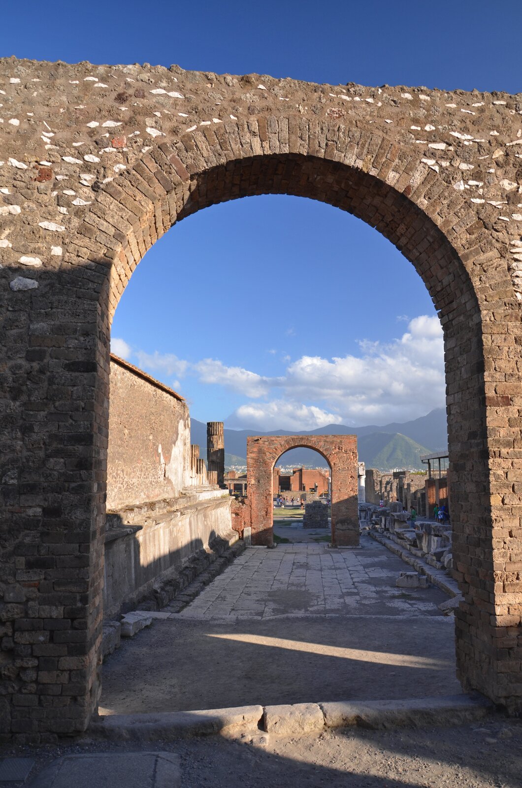 Ilustracja przedstawia widok na ruiny miasta Pompeje. Na zdjęciu znajdują się kamienne łuki i ściany budynków. Pomiędzy ramionami łuków widoczna jest kamienna droga. W tle widoczne są góry.
