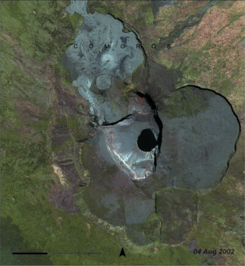 Zdjęcie z 2002 roku przedstawia wulkan na Komorach z lotu ptaka. Na obrazku widoczny zielono – brązowy teren dookoła wulkanu, szare zbocza oblane zastygłą lawą oraz pośrodku duży czarny krater. 