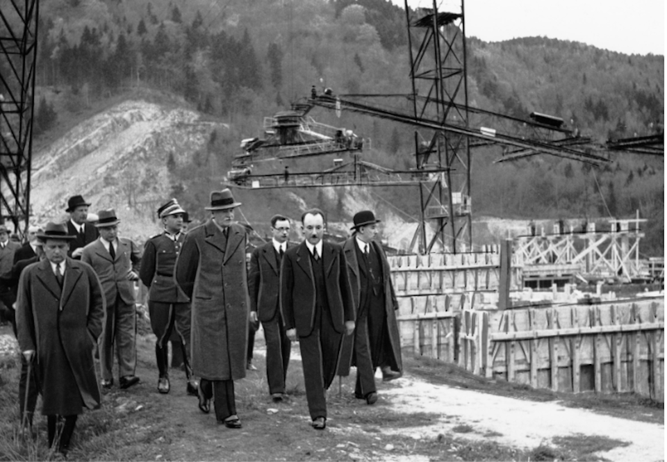 Ilustracja przedstawia grupę mężczyzn elegancko ubranych  idących górską drogą obok znajdującej się po ich prawej stronie dużej budowy. Z tyłu za mężczyznami widać gęsty las. 